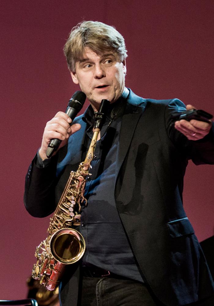 Jörgen Pettersson talar i en mikrofon med sin saxofon i en rem runt halsen